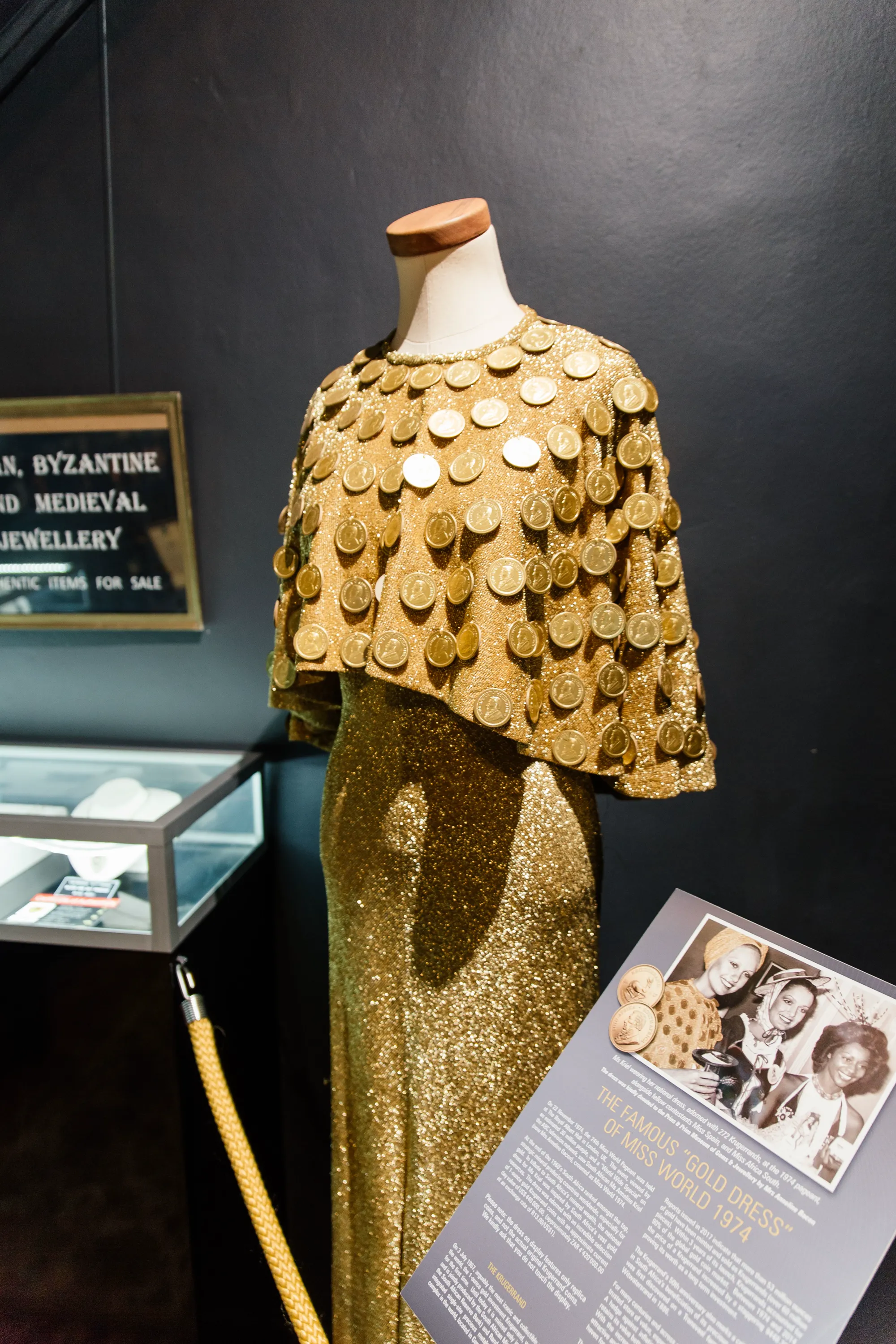 The Miss World 1974 Krugerrand gown on display in the museum, adorned with 272 replica Krugerrand coins, with the archival photograph of Anneline Kriel and the story plaque beside it
