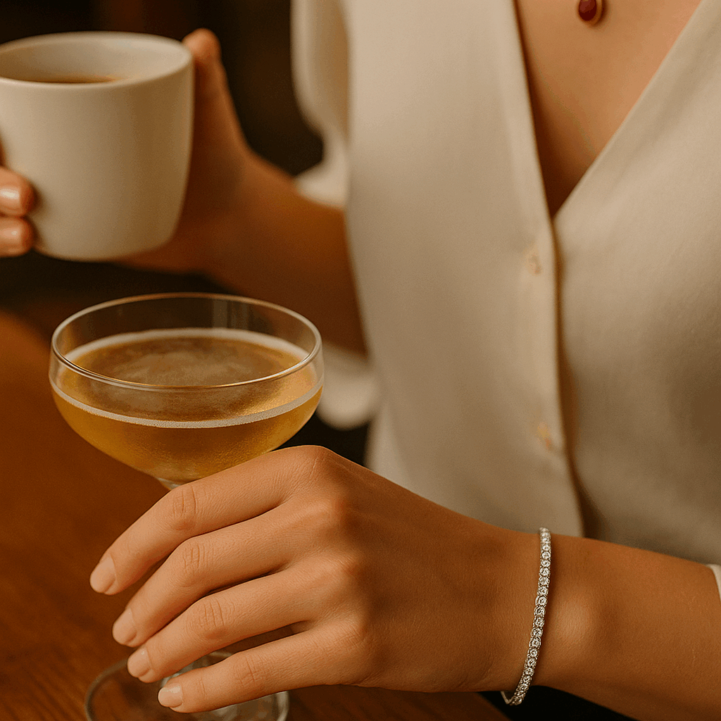 A woman transitioning from day to night, holding a coffee cup in one hand and a cocktail glass in the other, wearing a diamond tennis bracelet and ruby pendant, illustrating wearable luxury.